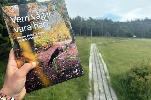 Hand håller omslaget till antologin Vem vågar vara här? i friluftsmiljö.